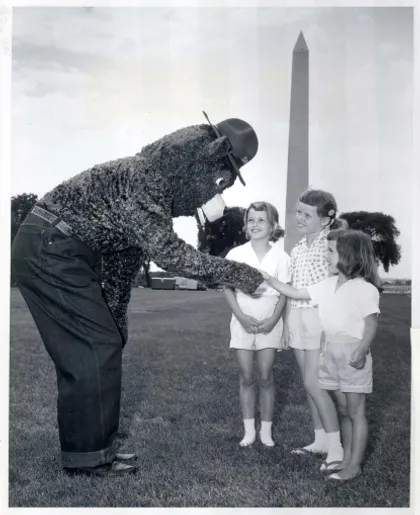 Una persona vestida con un disfraz del oso Smokey  interactuando con tres niños en una zona cubierta de césped, con el Monumento a Washington visible al fondo.