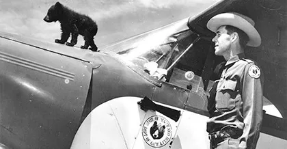 El cachorro Smokey sobre la parte delantera de un avión, con Ray Bell parado a su lado observándolo.